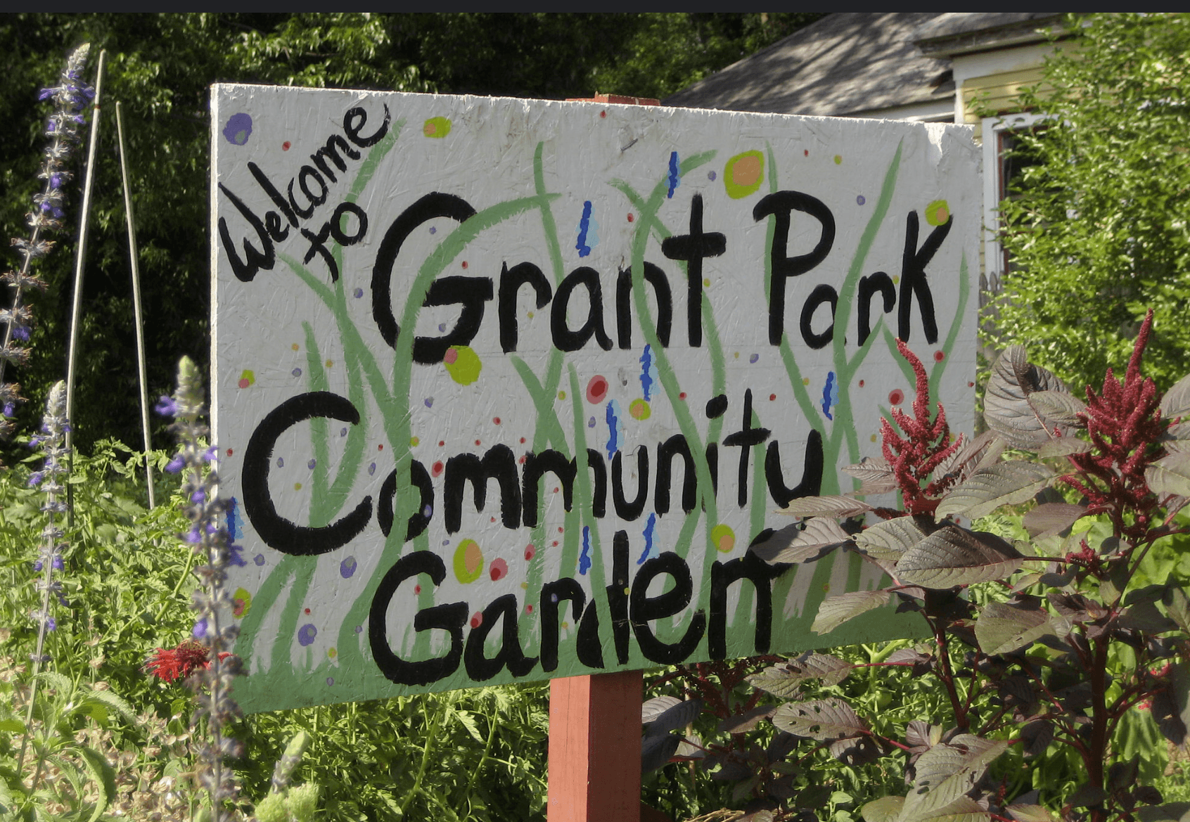 A pretty, hand-painted sign welcomes everyone to a community garden full of flowering plants.