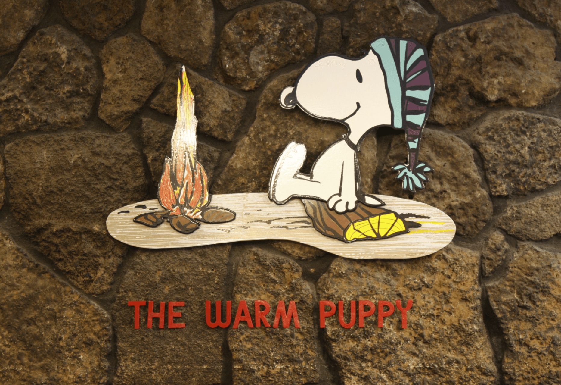 The Peanuts Cartoon character Snoopy is shown wearing a stocking hat and warming himself at a cozy campfire.