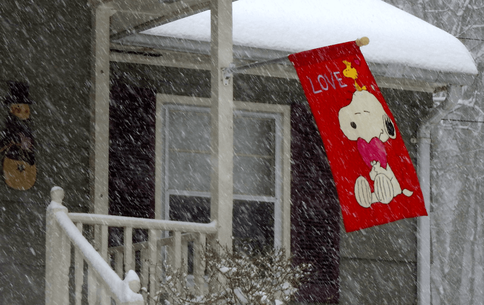 A cheerful banner with the Peanuts cartoon character Snoopy and the word "Love" sways from the porch of a house in a winter snow storm.